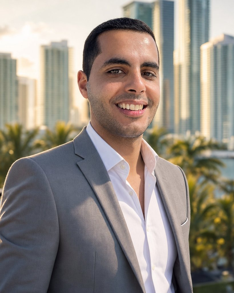 Mark Elbadramany in a light grey suit on a terrace, with Miami waterfront high-rises and palm trees behind him.